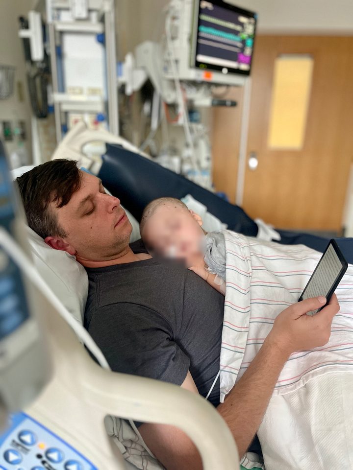 A father resting in a hospital bed with his young daughter asleep on his chest after her surgery; her face is softened for privacy